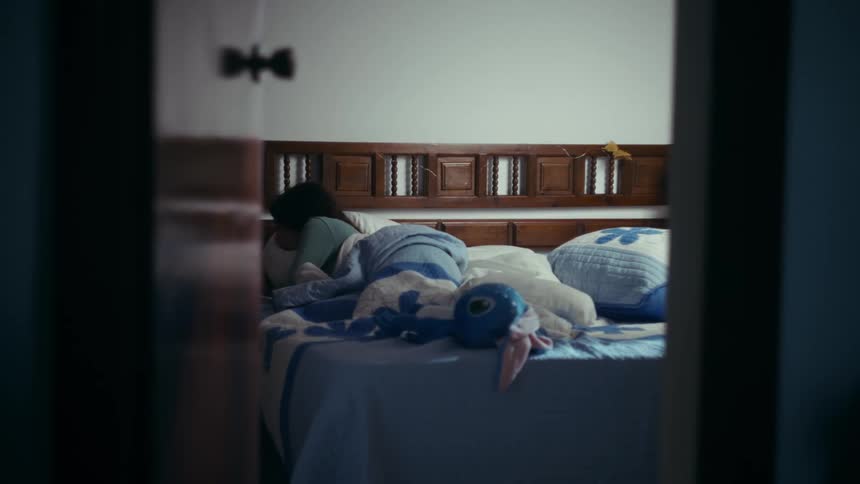 A woman lies in bed under a blanket waking up viewed from a doorway