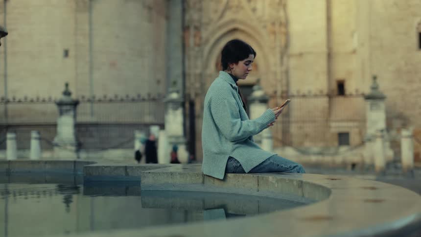 A woman sits by a fountain on a video call visibly emotional and crying