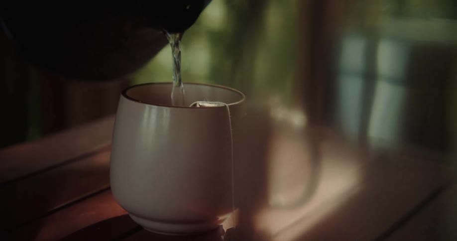 Hot water is poured from a dark kettle into a lightcolored mug which contains a tea bag the focus is on the steady stream of water filling the cup