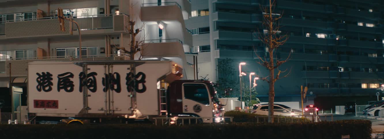 A truck with large japanese characters on its side is driving on a quiet city street at night with buildings in the background
