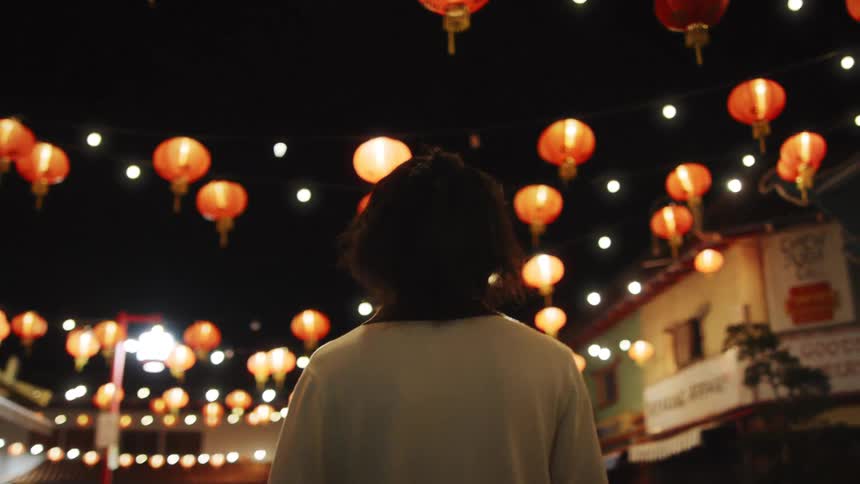 A young man walks through a lanternlit street at night surrounded by festive hanging lights