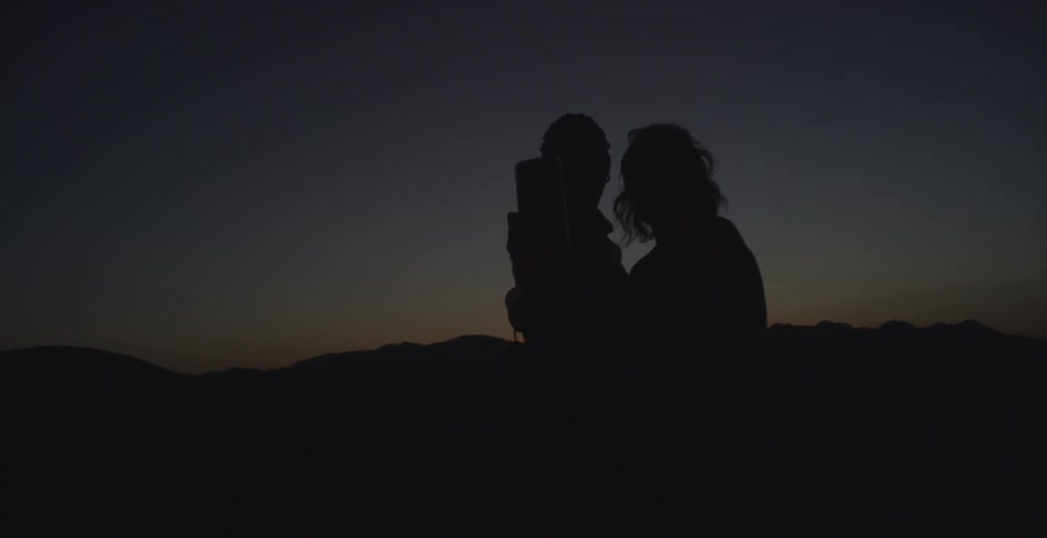 Two people take a picture with a phone silhouetted against a dim sunset sky and distant mountain ridges
