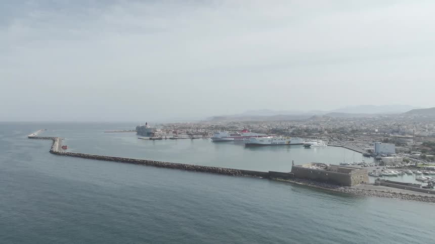 A city skyline is visible from the shoreline at midday with ships and boats docked in the bay
