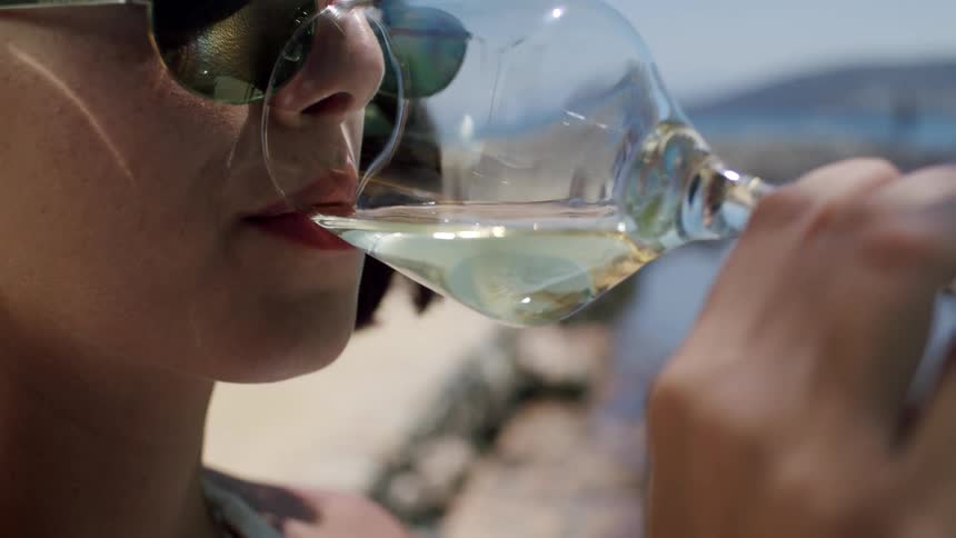 A woman is sitting by the beachside at midday drinking wine from a glass