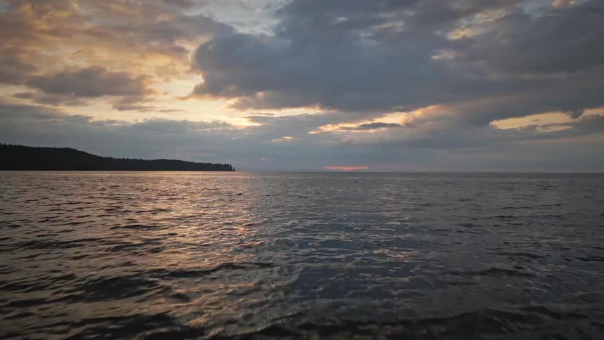 An aerial view flies backward over the ocean at sunset pulling back to reveal a dark rocky beach