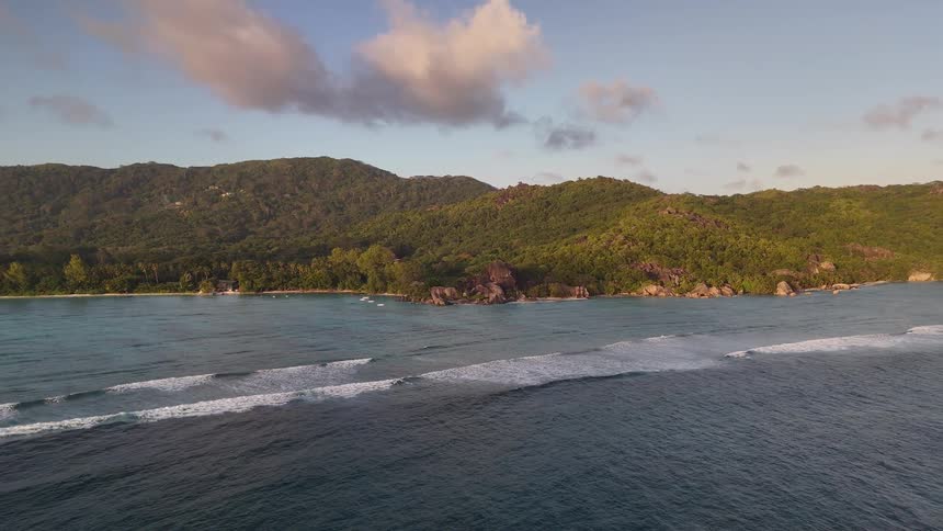 Gentle waves roll onto a sandy beach bordered by rocky outcrops and dense green hills under a partly cloudy sky