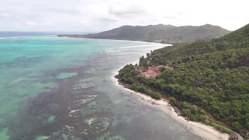 Gentle waves roll toward a lush forested coastline with rocky outcrops and clear turquoise waters