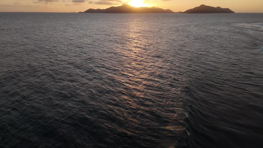 The sun sets behind a distant island casting golden light across calm ocean waters under a clear sky