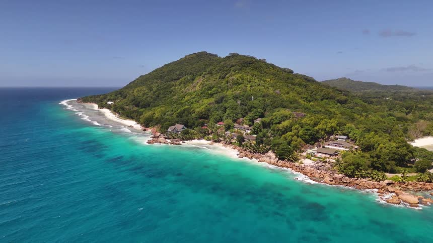 An aerial view lowers down moving closer to a tropical island dotted with houses and villas along its beautiful rocky coastline