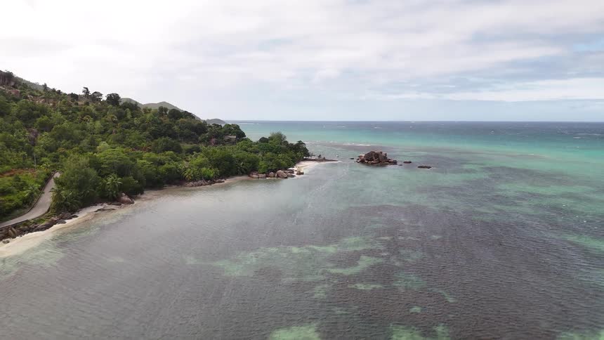Beginning near the sand the camera flies upwards and out over the water revealing a wide bay and a lush tropical coastline