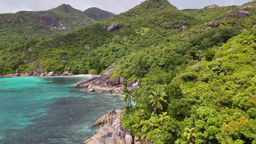 Turquoise waves lap against a rugged tropical coast lined with steep hills and rocky outcrops under a hazy sky