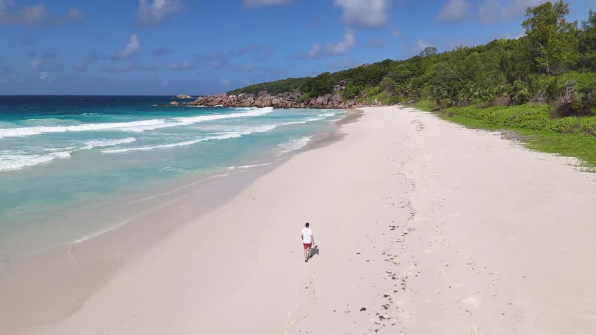 An aerial shot shows a person walking along a pristine white sandy beach next to a turquoise ocean with gentle waves the beach is bordered by lush green foliage and the sky is bright blue with scattered white clouds