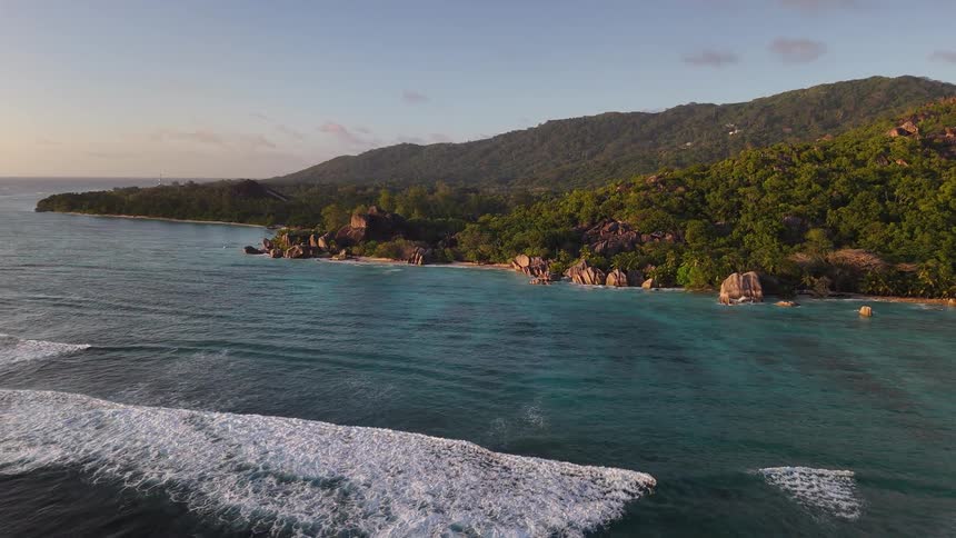 Gentle waves roll toward a lush rocky shoreline with dense forest and clear turquoise water