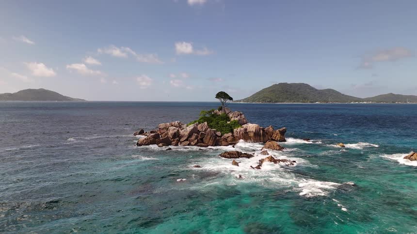 A small rocky island with a single windswept tree rises from turquoise ocean waves under a clear blue sky