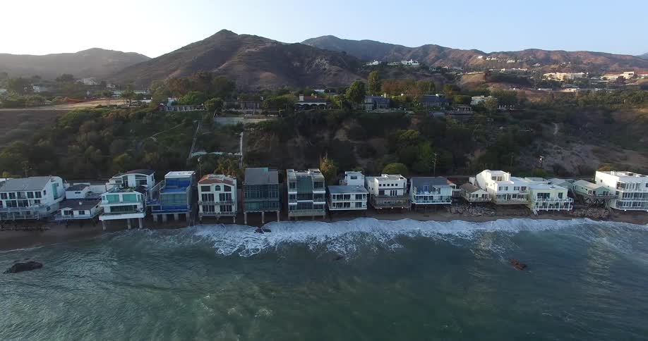 A row of beachfront houses in Malibu with waves crashing against the shore backed by a hillside with scattered trees and residential buildings