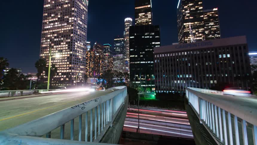 A timelapse shot tilts up from a graffiticovered railing to reveal a vibrant downtown Los Angeles skyline at night with traffic light streaks below