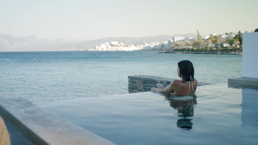 A woman is relaxing alone in an infinity pool overlooking a coastal town and the sea