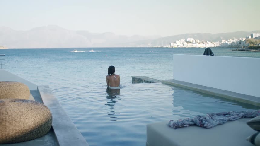 Woman enjoys vacation in infinity pool overlooking sea and town