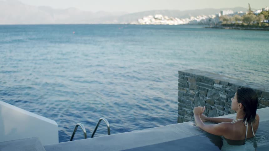 A woman relaxes in a pool overlooking a calm coastal town and the sea