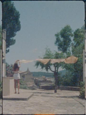 A woman hangs sheets on a line in a quaint courtyard overlooking distant hills