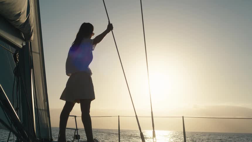 A woman is standing on the deck of a sailboat or yacht holding onto the rigging silhouetted against a bright sunset or sunrise over the ocean the scene is characterized by warm intense light and a calm sea