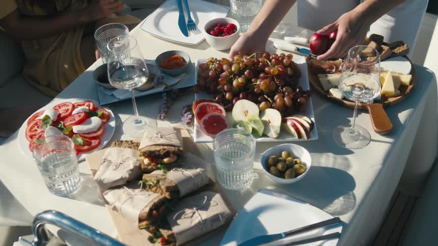 A sunlit table overflows with fresh fruits sandwiches and wine as hands reach to serve