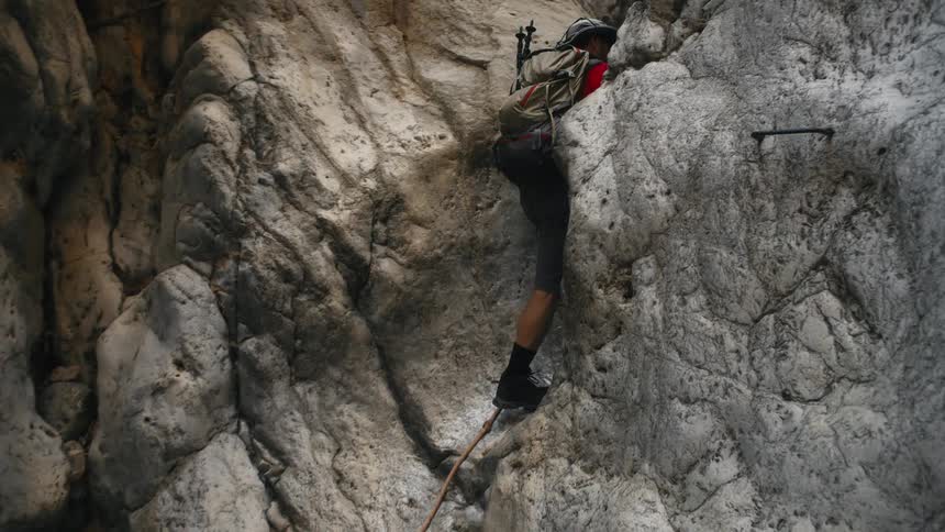Man wearing a hat and backpack using a rope climbing rocks between mountains