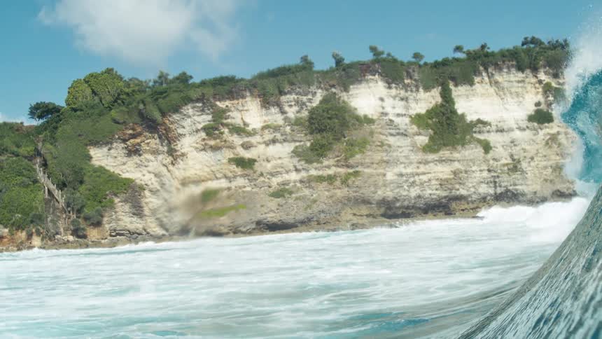 A powerful wave crests dramatically against a rugged cliffside backdrop under a bright blue sky