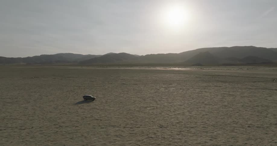 An aerial view shows a lone black car driving across a vast flat and arid desert landscape under a bright sunny sky distant mountains frame the horizon emphasizing the expansive and empty nature of the terrain