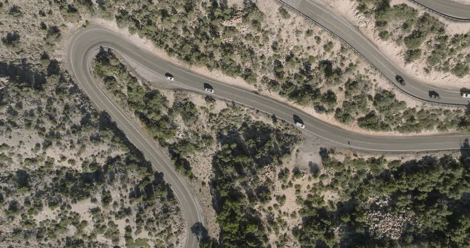 Vehicles drive along a winding asphalt road through a dry rocky desert with sparse shrubs and pale grass