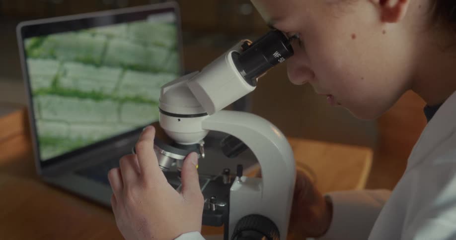 The girl is using a microscope as the display on her laptop shows an image