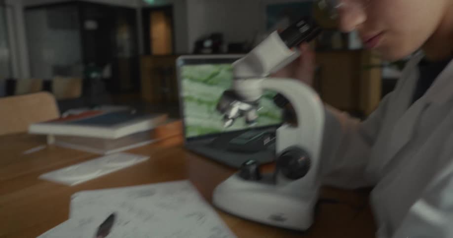 Young woman in lab coat writing and interacting with microscope