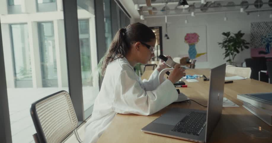 Young female scientist make notes while viewing a sample with a microscope at a table in a lab during daytime