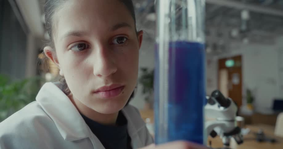 Young female scientist inspects a blue liquid in a cylindrical beaker in a lab during daytime