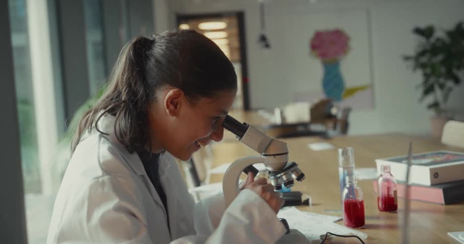 Happylooking teenager sitting in a white coat and using a microscope
