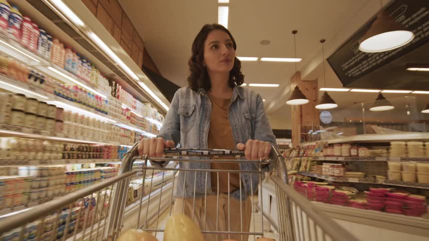 A woman pushes a shopping cart down an aisle in a well lit grocery store