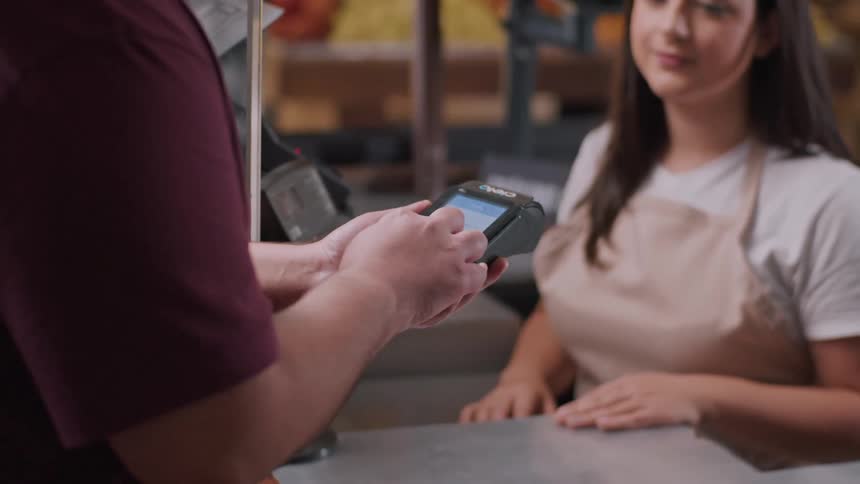 A man and a woman are seen at a checkout counter with the man holding a payment device the woman a cashier wearing an apron is looking at him with a slight smile