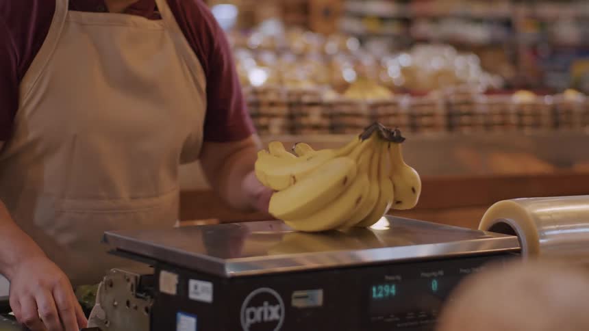 A transaction at a grocery store a woman hands a bunch of bananas to a person wearing an apron presumably a store employee the employee then places the bananas on a digital scale to weigh them