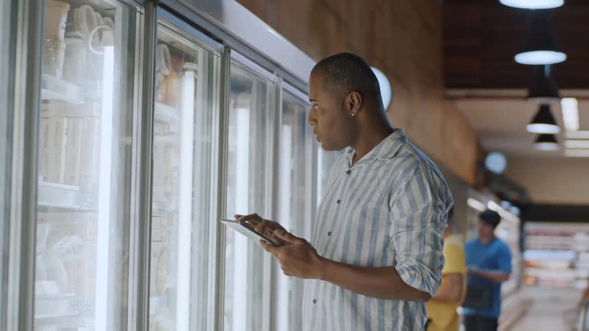 A man is holding a tablet and taking stock of items inside a refrigerator in a supermarket