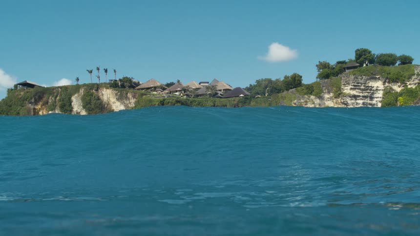 A beautiful blue ocean with the waterline visible in the background a lush green landmass with what appears to be buildings or structures emerges from the horizon under a clear blue sky the camera seems to be slightly submerged