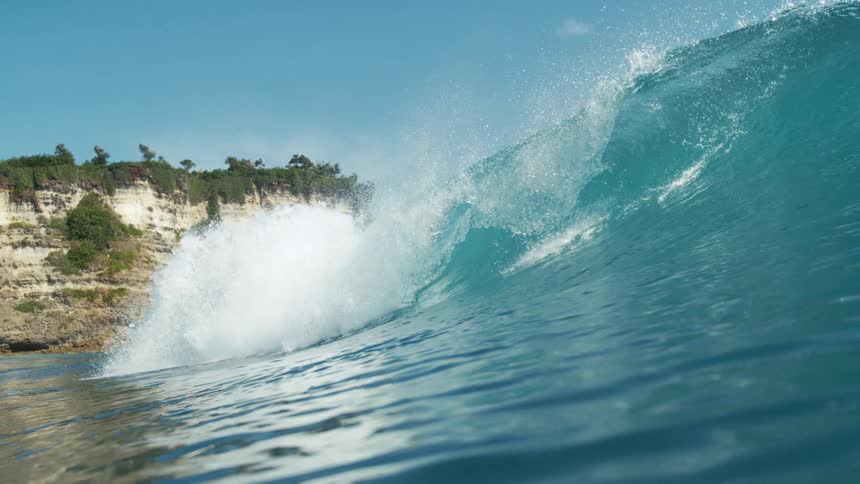 A powerful ocean wave rises near a rocky coastal cliff under a clear blue sky