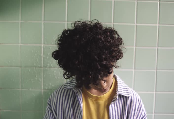 A teenage boy stands against a bathroom wall looking down at the floor