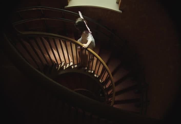 A highangle shot of a spiral staircase with a wooden railing and dark steps a person appearing to be a child is seen briefly ascending and descending the stairs