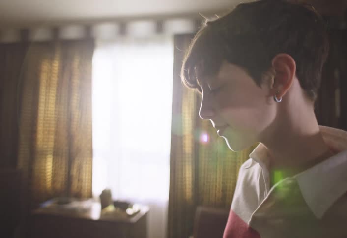 A young girl with short dark hair and wearing a striped shirt stands in a room with patterned curtains operating a vacuum cleaner sunlight streams in from a window on the left illuminating part of the room
