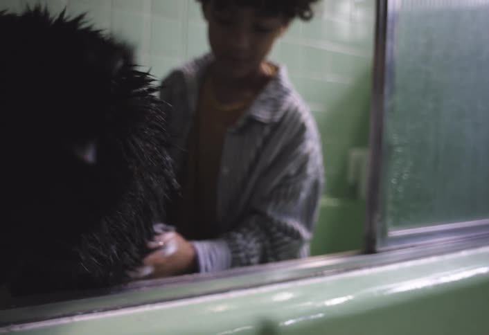 A boy with curly hair and a striped shirt is seen inside a bathroom with green tiled walls gently bathing a black dog in a tub he is focused on washing the dog creating a heartwarming scene of pet care