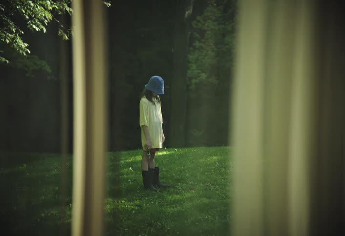 A child wearing a yellow dress and a blue bucket hat is standing outdoors in a lush green lawn the scene is viewed through a window with curtains creating a framed look at the child who is looking upwards seemingly at the sky or something above