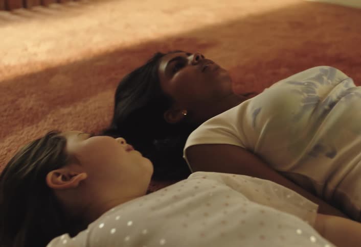 A young woman with dark long hair wearing a white tshirt lying on a red rug she is interacting with a younger child who is also on the floor smiling and appearing to be playing