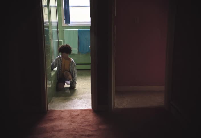 A person with curly hair wearing a striped shirt and a yellow shirt underneath is seen kneeling in a bathroom with light green tiled walls they are scrubbing the base of a toilet