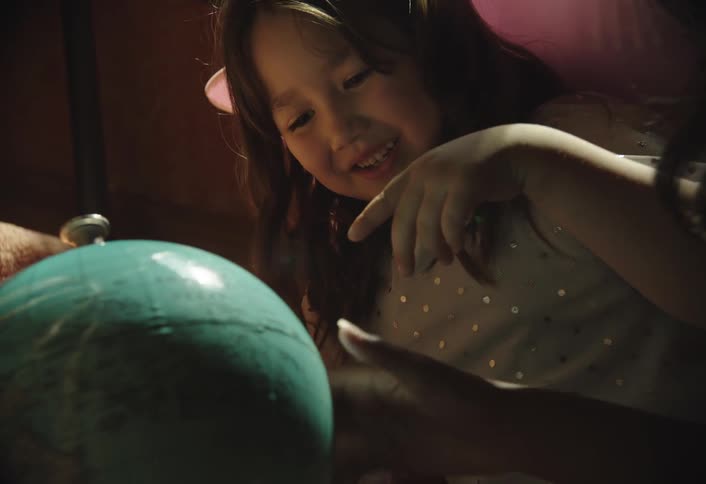 A child with long dark hair is seen closely examining a tealcolored globe turning it with their hands they appear to be engaged in learning or exploration with a slight smile on their face
