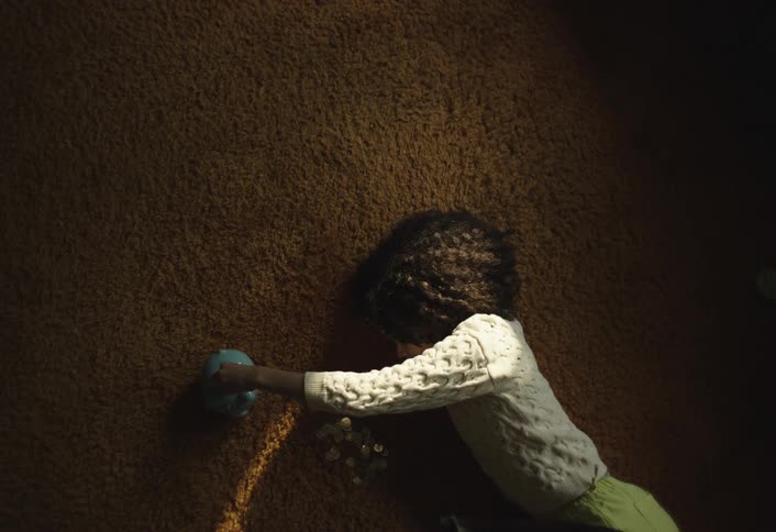 A young person with curly dark hair is lying on a brown carpet reaching for a light blue piggy bank there are coins scattered on the carpet near the piggy bank illuminated by a beam of sunlight